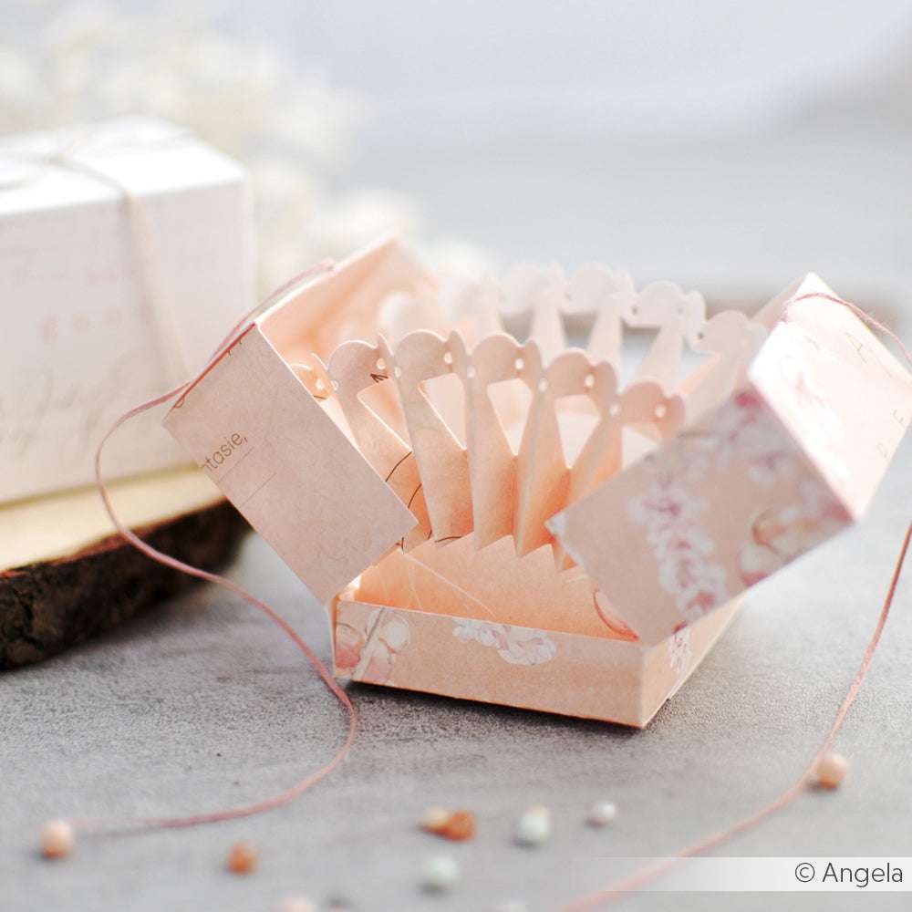 Eine dekorative rosafarbene Geschenkbox von Alexandra Renke mit einem aufwendigen Pop-up-Design, das mit DieCut-Systemen hergestellt wurde, liegt offen auf einer grauen Oberfläche.