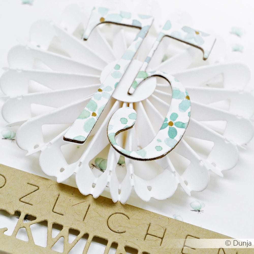 Die Alexandra Renke Motivstanze Rosette 4 wurde verwendet, um eine weiße Papierrosette mit einer floralen Zahl 75 zu gestalten, die neben einem Banner aus Kraftpapier steht.