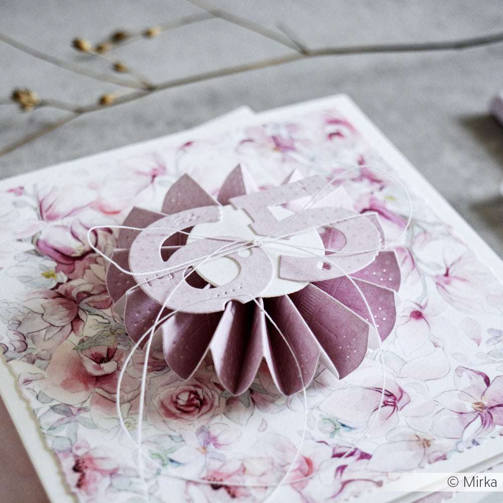 Eine rosafarbene 3D-Blumendekoration aus Papier von Alexandra Renke, hergestellt mit Designpapieren und der Motivstanze Rosette 5 Stanzsysteme, hängt an einer weißen Schnur vor einem pastellfarbenen Blumenkartenhintergrund.
