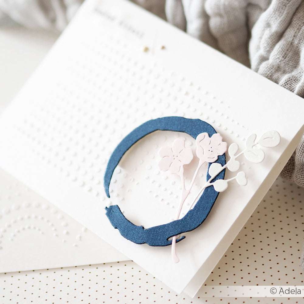 Weiße Karte mit geprägten Punkten, einem blauen Kreis, der mit der Alexandra Renke Motivstanze Maru Kreis ausgestanzt wurde, und zarten rosafarbenen und weißen Alexandra Renke Designpapieren mit Blumenausschnitten, die darauf gelegt wurden.
