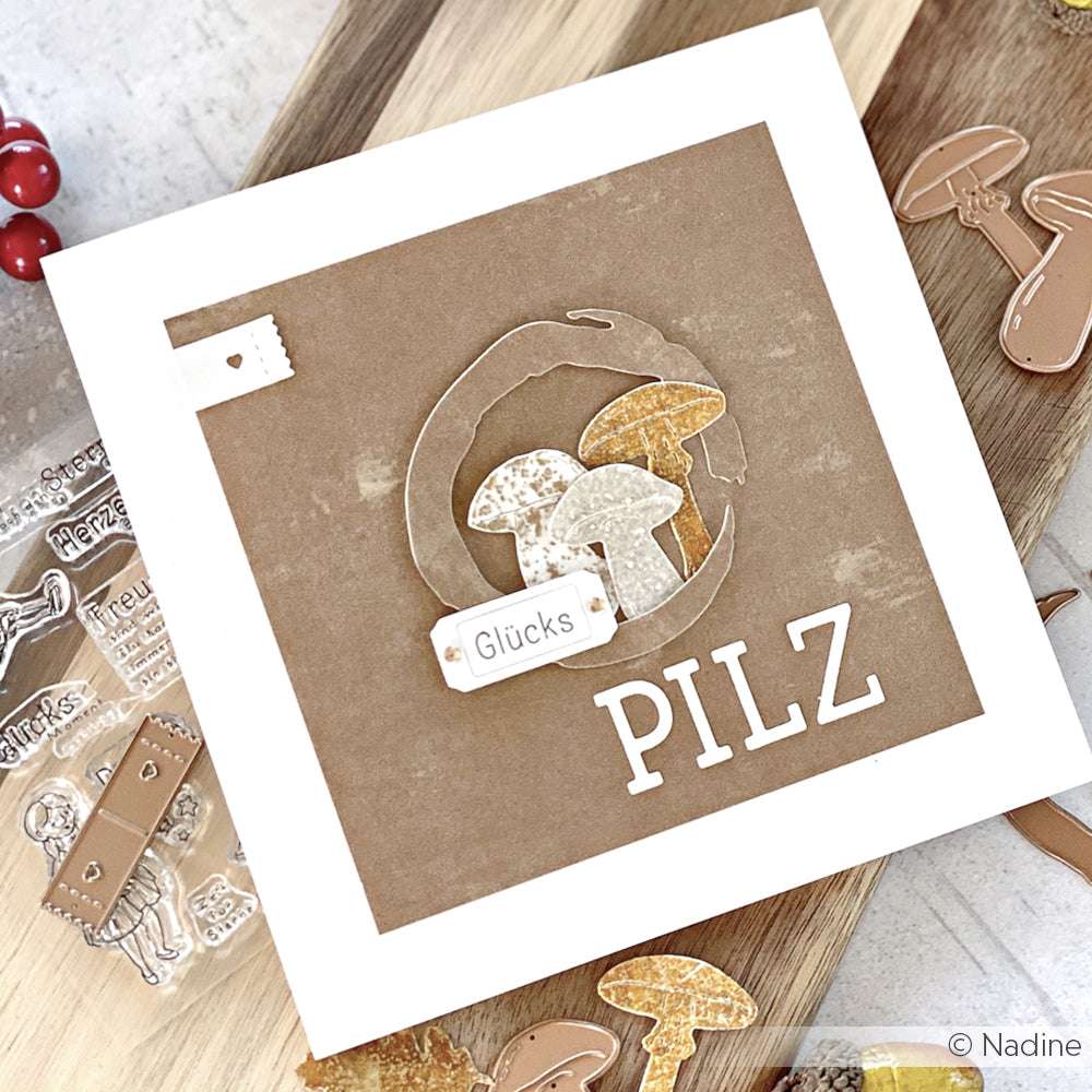 Quadratische Karte von Alexandra Renke mit Pilzausschnitten und den Wörtern "Glücks" und "PILZ" auf braunem Hintergrund, mit Bastelmaterial und Motivstanze Maru Kreis Dies in der Nähe.