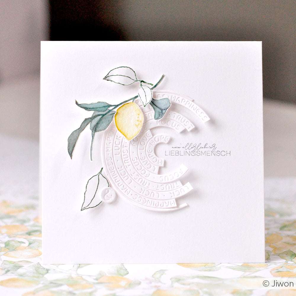 Weiße Karte von Alexandra Renke mit geprägtem kreisförmigem Text - Motivstanze Wortkreise - mit einer aquarellierten Zitrone und Blättern oben links, ideal für kreative Projekte mit Stanzsystemen.