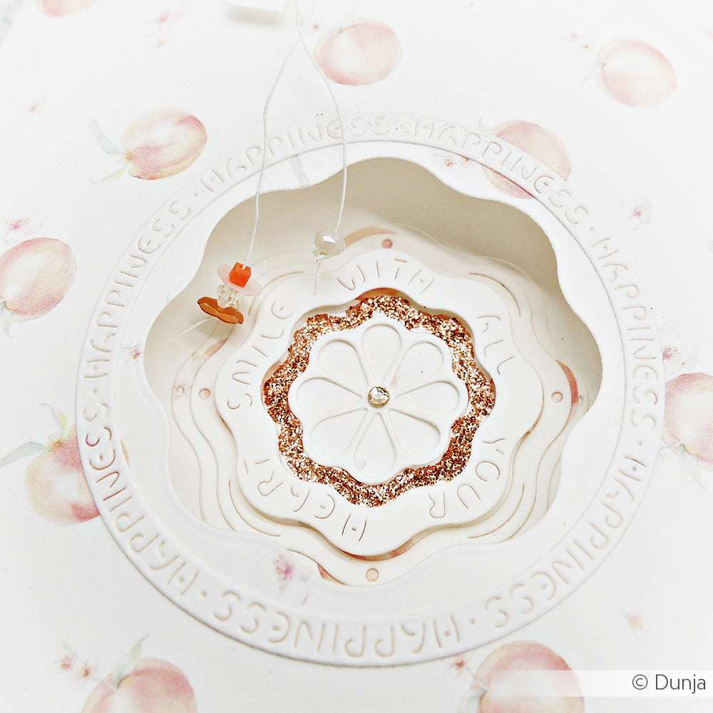 Eine runde, verzierte Karte von Alexandra Renke mit pfirsichfarbenen Illustrationen, geschichteten Blumenausschnitten aus Designpapier, Glitter, einem Schmetterlingsanhänger und einem eleganten "Motivstanze Rahmen Smile" Akzent.