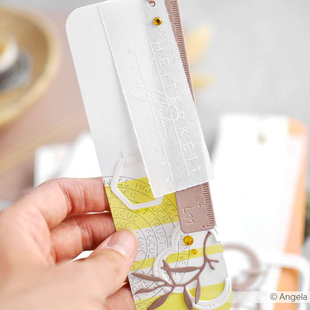 Eine Hand hält ein dekoratives Lesezeichen von Alexandra Renke, das mit Designpapier hergestellt wurde und gelbe, weiße und goldene Muster sowie geschichtete Papierverzierungen aufweist.