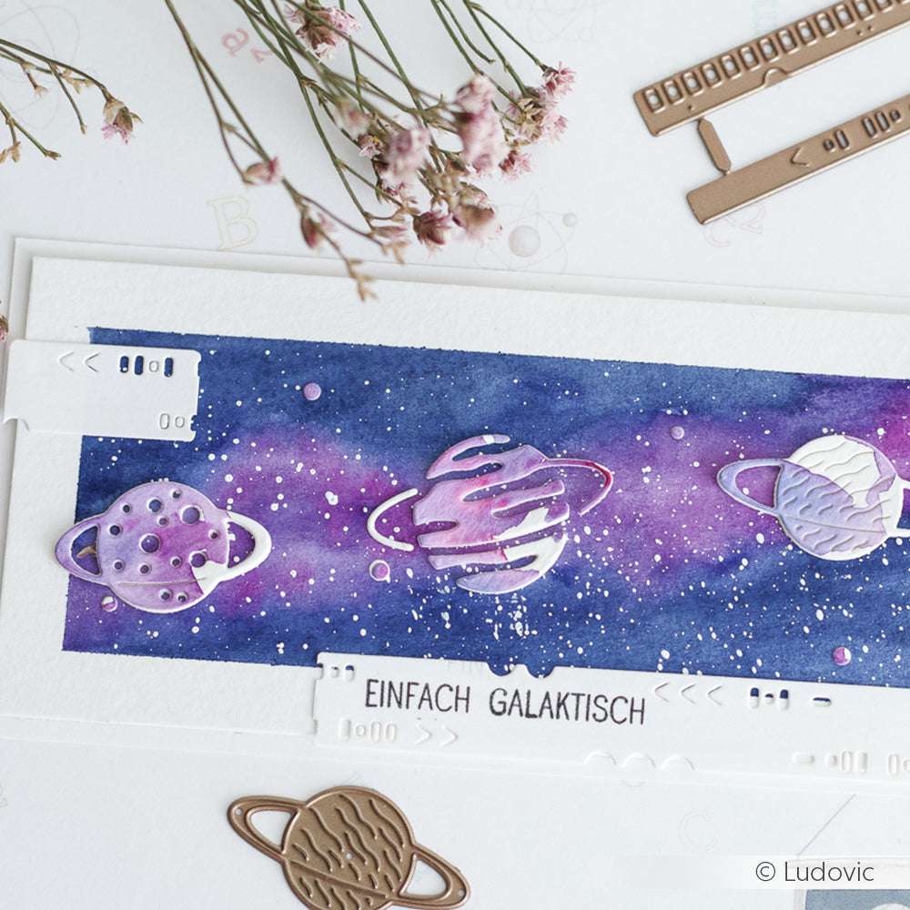 Eine aquarellierte Galaxie mit metallischen Planetenausschnitten, hergestellt mit Alexandra Renke Designpapieren und Motivstanze 3 Planeten, beschriftet mit "EINFACH GALAKTISCH", umgeben von Blumen und Bastelwerkzeug.