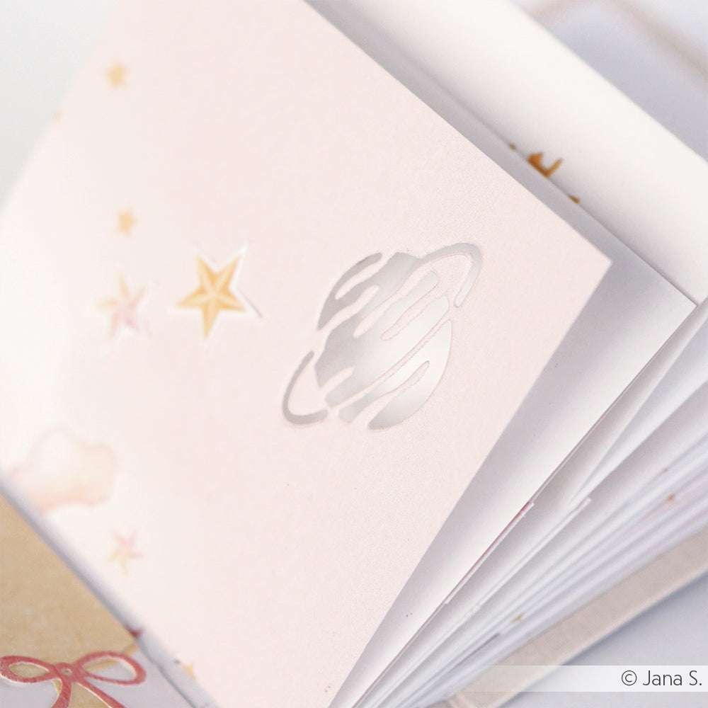 Aufgeschlagenes Notizbuch mit einer blassrosa Seite mit Planeten- und Sternenausschnitten, die mit Alexandra Renke Designpapieren und dem Motivstanze 3 Planeten DieCut System hergestellt wurden.