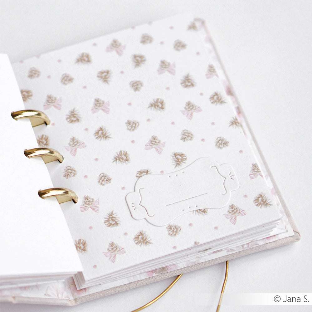 Offener Planer mit goldenen Ringen und einem pastellfarbenen, floralen Einband von Alexandra Renke, auf einer weißen Fläche mit eleganten Designpapieren und der Motivstanze Etikett Vic 5 für kreative Planung.