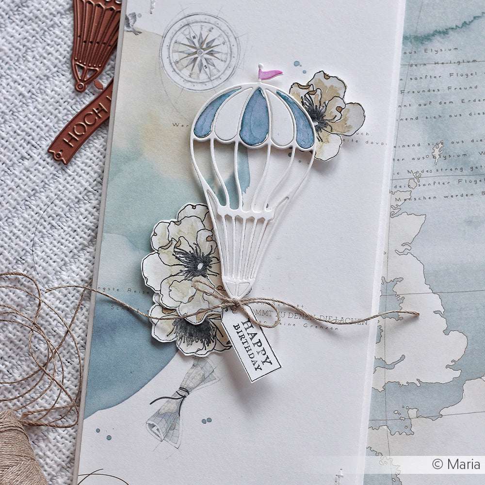 Handgemachte Karte mit einem blauen Heißluftballon, gestaltet mit der Alexandra Renke Motivstanze Heißluftballon, floralen Akzenten, Designpapieren, Garnschleife und einem "Happy Birthday"-Anhänger auf strukturiertem Papier.