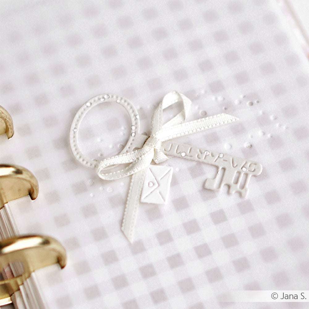 Eine weiße Schlüssel- und Schleifenverzierung, erstellt mit Alexandra Renke DieCut Systeme Motivstanze Schlüssel, ruht auf einem hellen, karierten Hintergrund neben einer Spiralheftung.