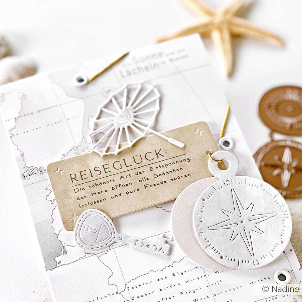 Eine Karte zum Thema Reisen von Alexandra Renke zeigt gestanztes Papier - einen Kompass, einen Regenschirm, eine Karte und einen REISEGLÜCK-Anhänger - die mit Stanzen aus Stahl präzise hergestellt und mit einem Motivstanze-Schlüssel für zusätzliches Abenteuer versehen wurden.