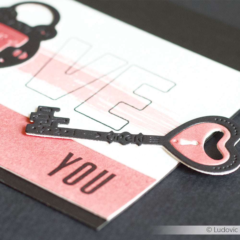 Ein dekorativer Schlüssel aus der Alexandra Renke Motivstanze Schlüssel hat eine herzförmige Spitze und ist auf einer Karte mit der Aufschrift LOVE YOU in Pink und Schwarz angebracht.