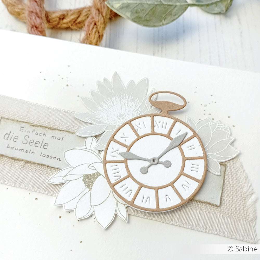 Handgemachte Karte von Alexandra Renke mit Designpapier, einer gestanzten Taschenuhr, Blumen und deutschem Text auf einem strukturierten Bandhintergrund.