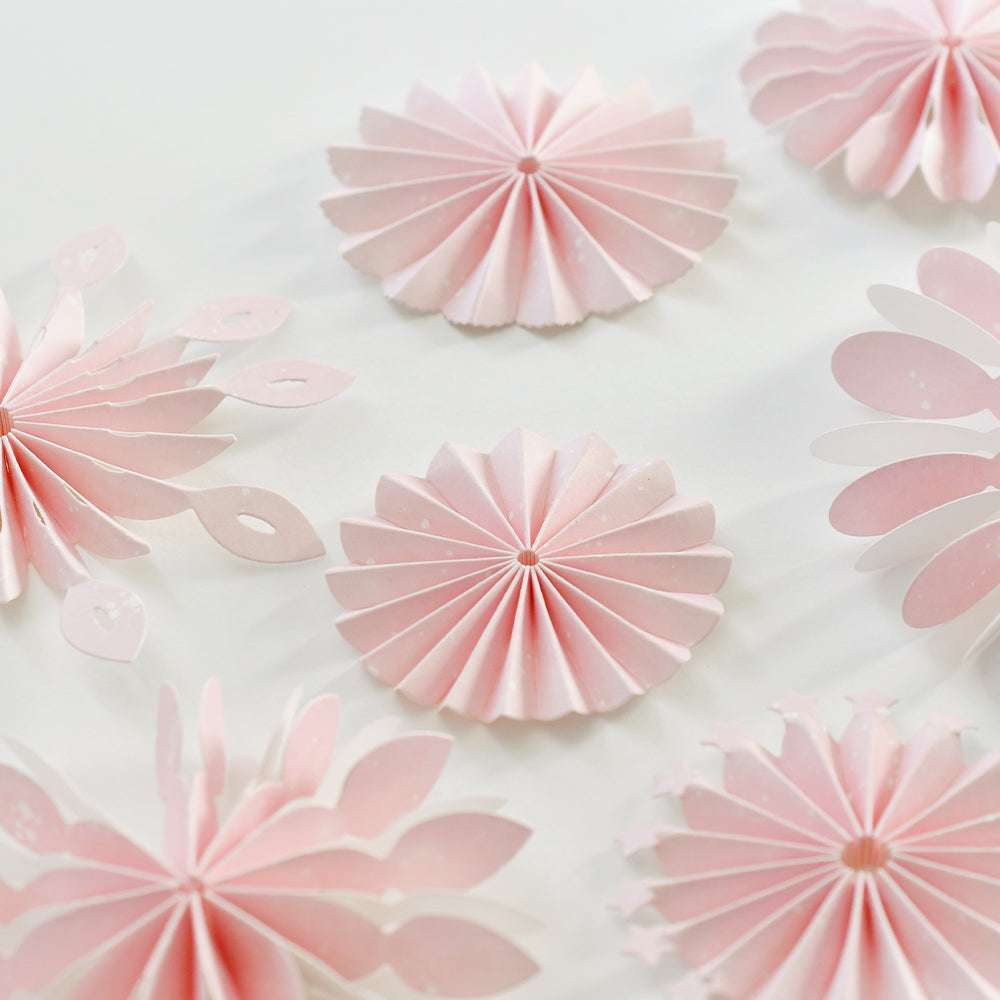 Rosafarbene Papierblumen mit Falten- und Blütenmotiven, die mit der Alexandra Renke Motivstanze Rosette Basic Stanzen gefertigt wurden, sind kunstvoll auf weißem Hintergrund arrangiert.