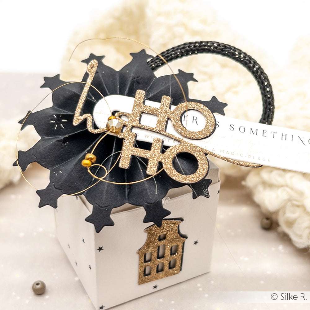 Eine festliche Geschenkbox von Alexandra Renke mit schwarzem Sterntopper, goldenen Ho-Ho-Ho-Buchstaben, dekorativen Designpapier-Akzenten und einem kleinen Hausornament.