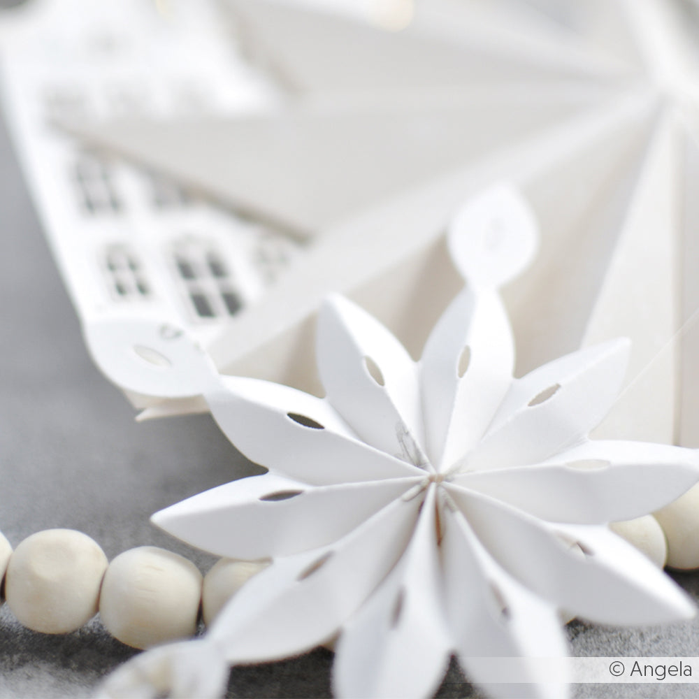 Nahaufnahme eines Schneeflockenornaments aus weißem Papier, hergestellt mit der Alexandra Renke Rosette Schneeflocke Stanze, mit Holzperlen und einem verschwommenen weißen Papierhaus im Hintergrund.