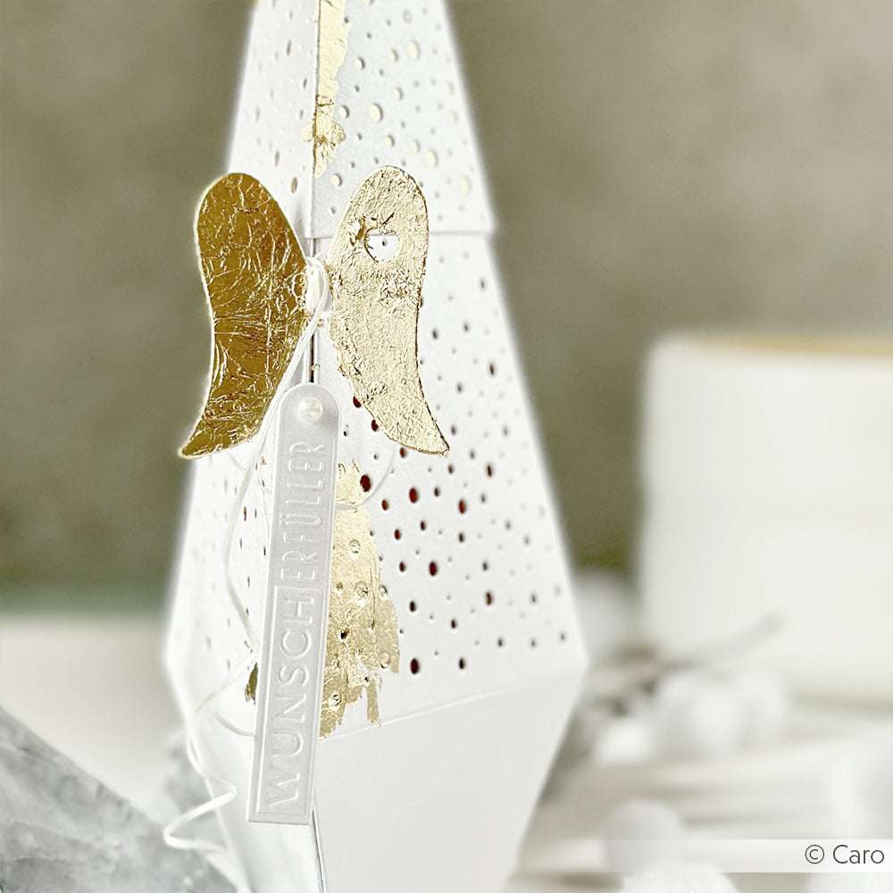 Weiße geometrische Schachtel mit goldenen Engelsflügeln, hergestellt mit den Engelsflügel-Stanzformen von Alexandra Renke, veredelt mit einem WUNSCHERFÜLLER-Anhänger und ausgestellt auf einem neutralen Hintergrund.
