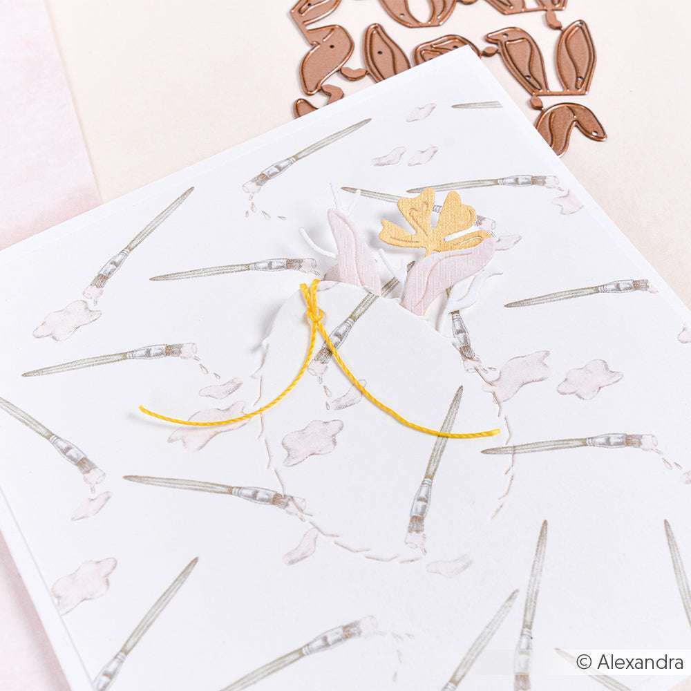 Eine weiße Karte von Alexandra Renke mit einem floralen Ausschnitt, der mit den Motivstanze Abrissrahmen Ei ausgestanzt wurde, verziert mit gelbem Garn und zarten rosa und goldenen Blumenakzenten.