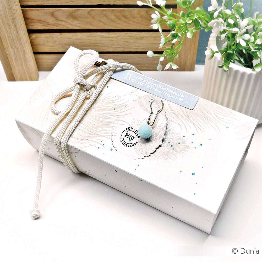 Eine weiße Alexandra Renke Geschenkbox mit blauem Bommel-Schlüsselanhänger und weißer Kordel steht neben einer kleinen Topfpflanze - ideal zum Verschenken von Stanzen für DieCut Systeme oder zum Ausstellen Ihrer Lieblingskreationen von Designpapiere 200g.