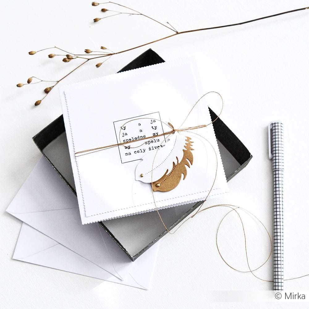 Eine quadratische weiße Karte von Alexandra Renke aus Designpapier mit einer goldenen Schnur und einem Federanhänger wird neben einer schwarzen Schachtel, einem Umschlag und einem Stift auf weißem Hintergrund gezeigt.