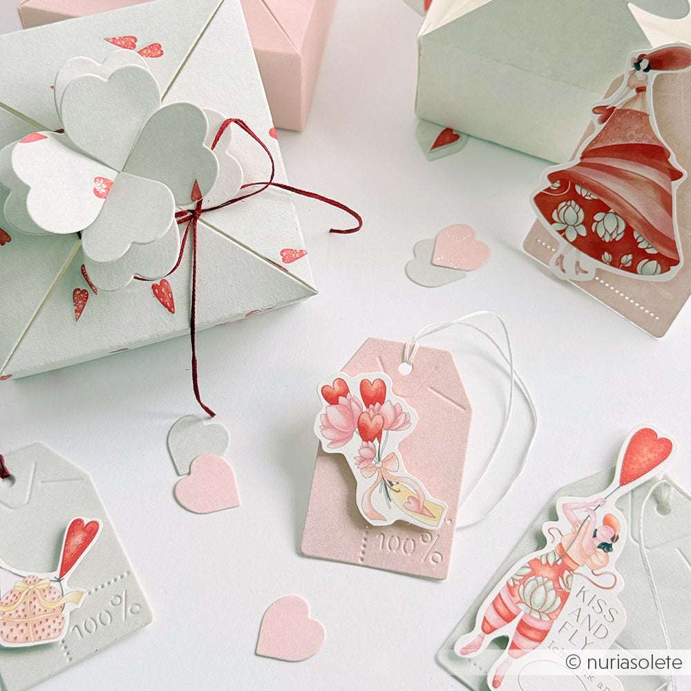 Pastellfarbene Geschenkschachteln und -anhänger, die mit Alexandra Renke Stanzen für Designpapier hergestellt und mit Herzen und charmanten Valentinsillustrationen verziert wurden, werden auf einer weißen Fläche präsentiert.