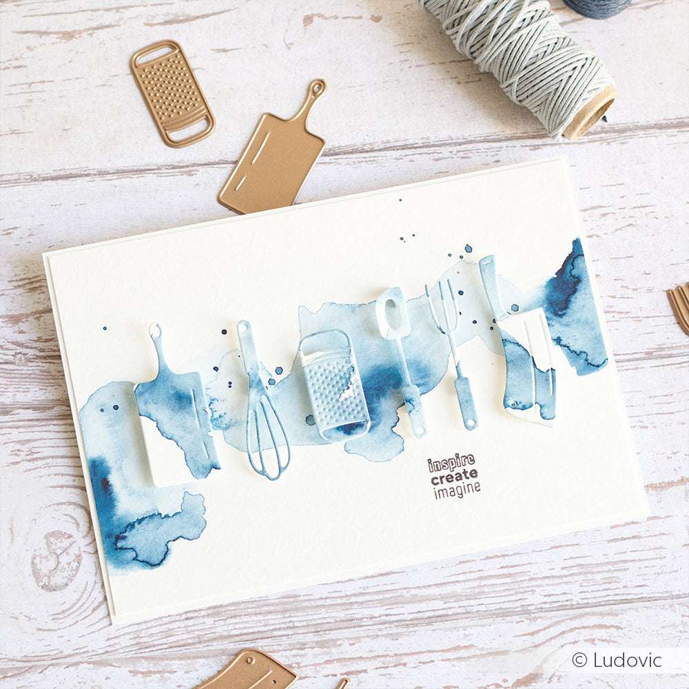 Handgefertigte Karte von Alexandra Renke mit blauen Aquarellspritzern und Küchenutensilien-Set Stanzungen von DieCut Systeme auf einer weißen Holzoberfläche.
