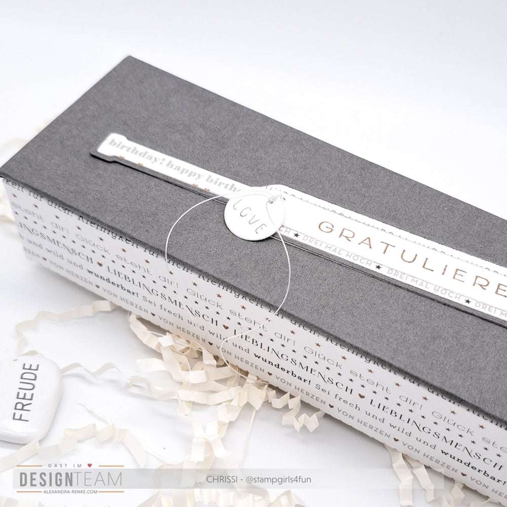 Graue Geschenkschachtel mit weißen Anhängern "LOVE" und "GRATULIERE", umgeben von weißem Knitterpapier - ideal, um Ihre Alexandra Renke Designpapiere zu präsentieren und mit der Motivstanze Die 'Lange Flaschenpost' kreative Akzente zu setzen.