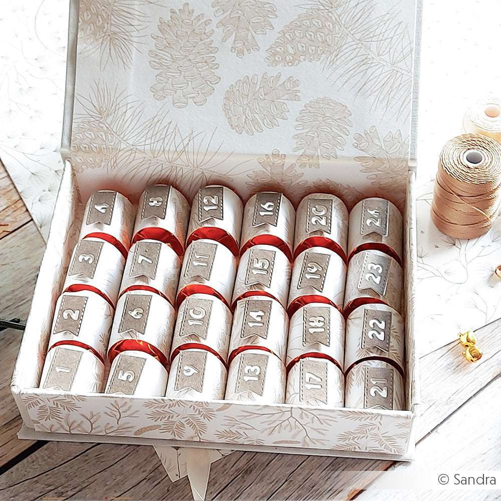 Eine dekorative Schachtel von Alexandra Renke aus Designpapier enthält 24 nummerierte, verpackte Leckereien, die in vier Reihen angeordnet sind - wie bei einem Adventskalender.