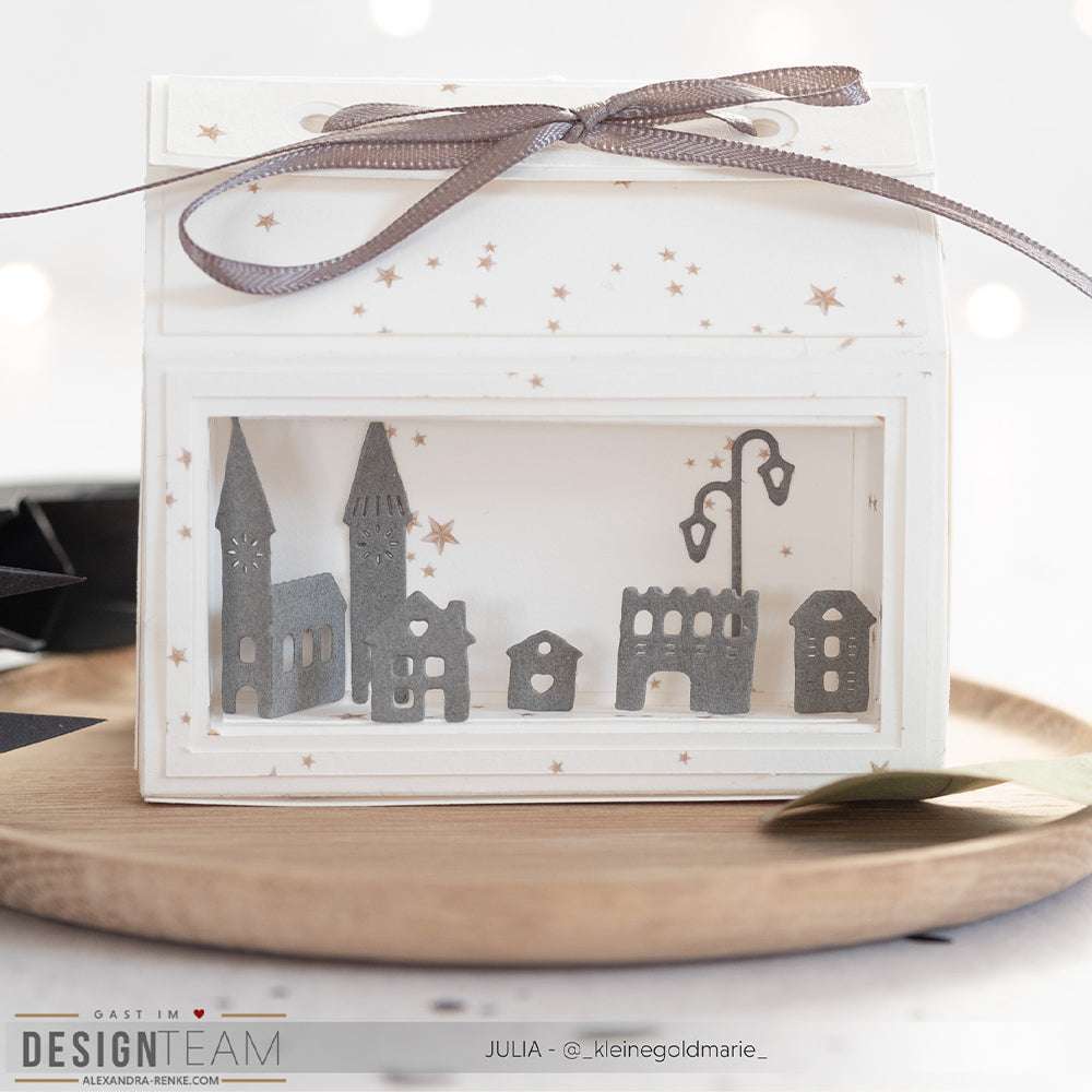 Ein weißer Geschenkkarton mit Klarsichtfenster beherbergt im Inneren ein graues Miniatur-Papierdorf, das mit der Kleinen Häuserreihe mit Kirche von Alexandra Renke und eleganten Designpapieren gestaltet wurde.
