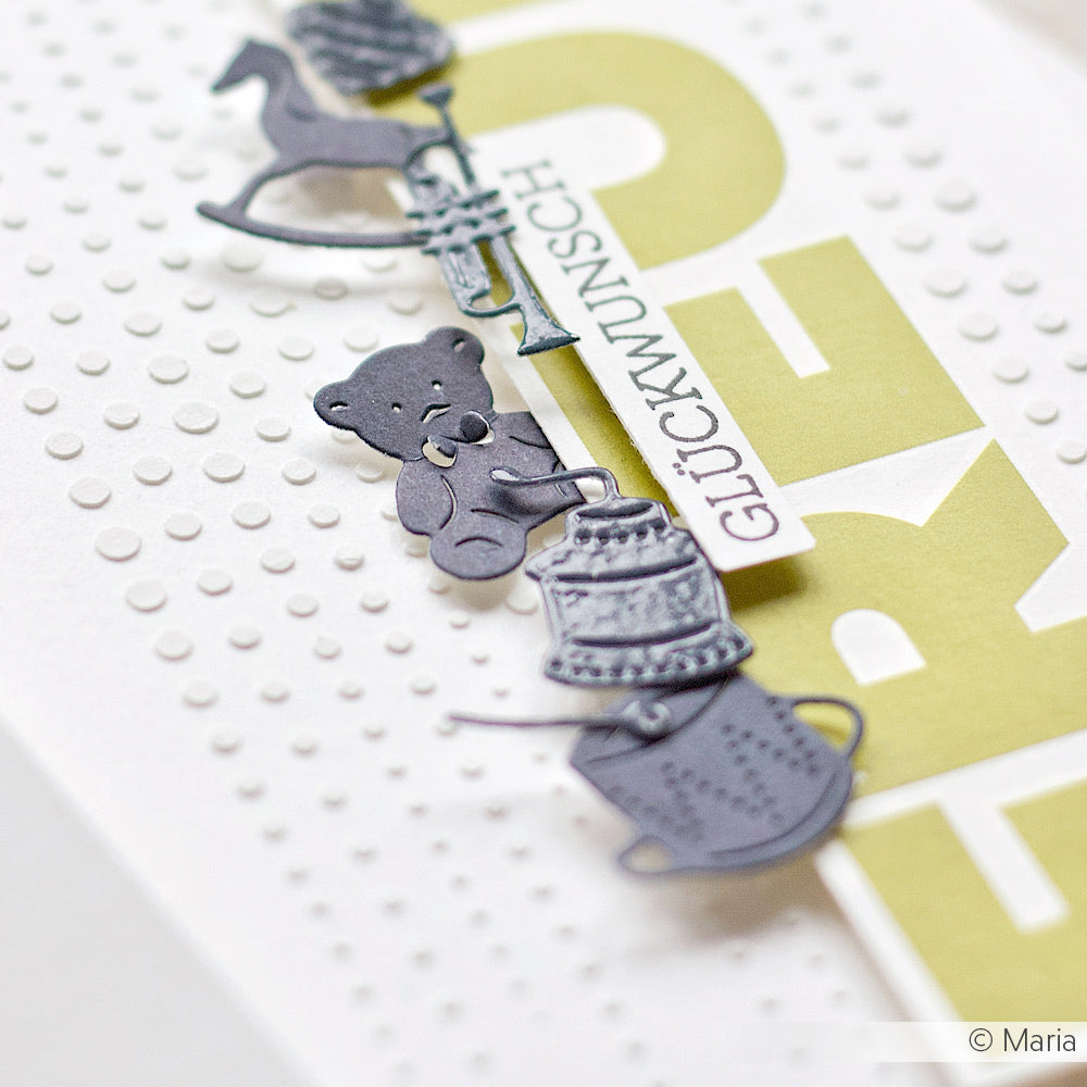 Auf einer gepunkteten weißen Karte mit grünem "GLÜCKWUNSCH"-Schriftzug liegen graue Papierausschnitte von Spielzeugen und einer Tasse, die mit den DieCut Systemen und der Motivstanze Vintage Spielzeug von Alexandra Renke hergestellt wurden.