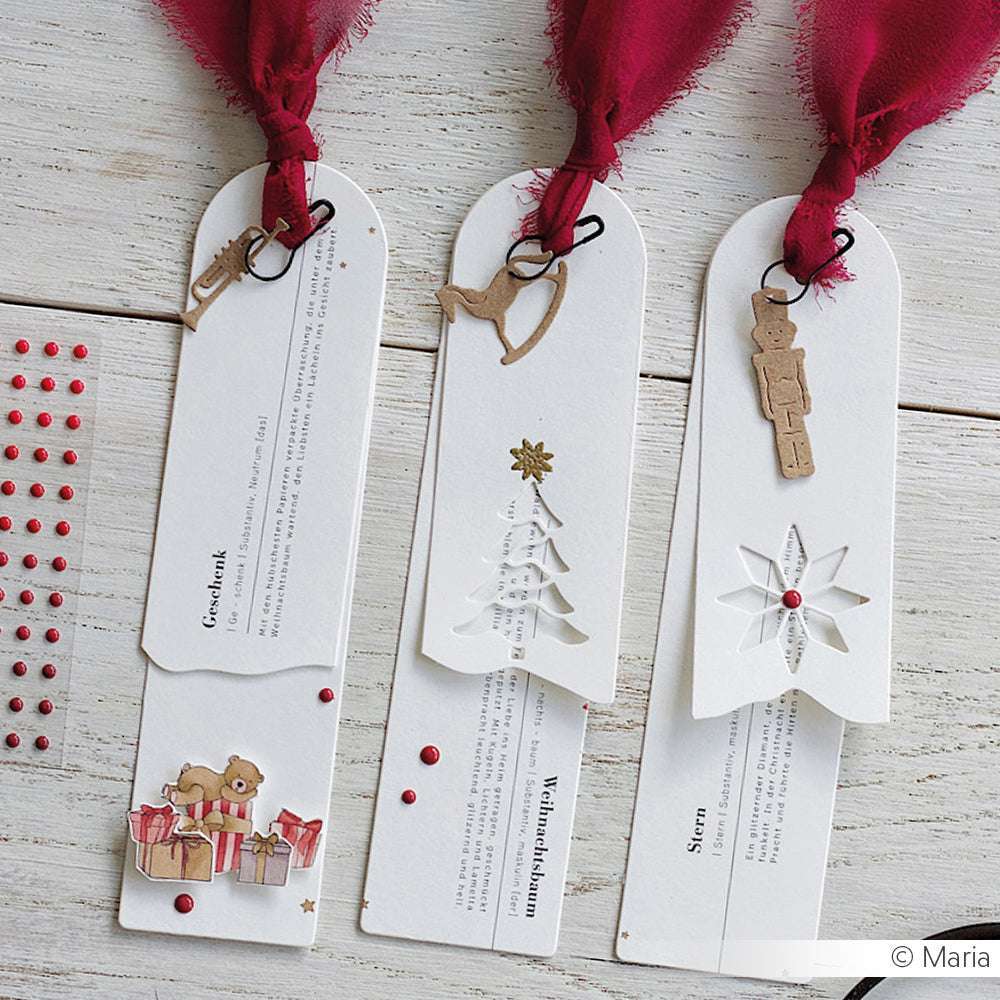Drei festliche weiße Lesezeichen mit roten Bändern, alten Weihnachtsanhängern und weihnachtlichen Dekorationen aus Alexandra Renke Stanzen für DieCut Systeme und 200g Designpapieren auf einer Holzoberfläche.