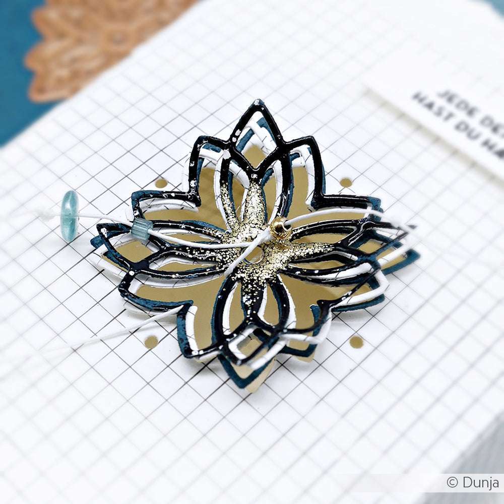 Eine metallische, geschichtete Blumenverzierung sitzt auf einem gerasterten Bogen von Alexandra Renke Designpapier, umgeben von Perlen und goldenen Akzenten.