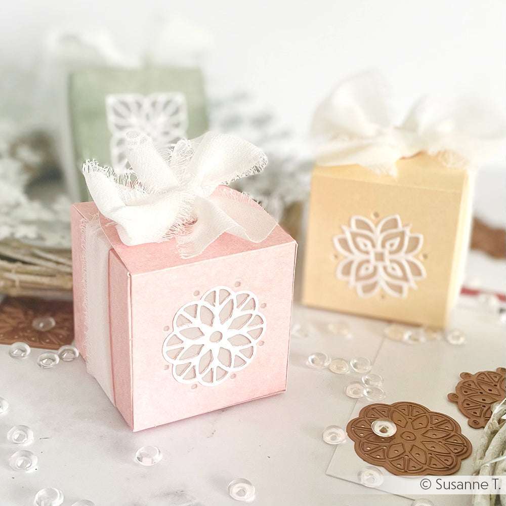 Drei pastellfarbene Geschenkschachteln mit Schleifenband und komplizierten Ausstanzungen, die mit Alexandra Renke Stanzen hergestellt wurden, umgeben von Dekorationsartikeln.