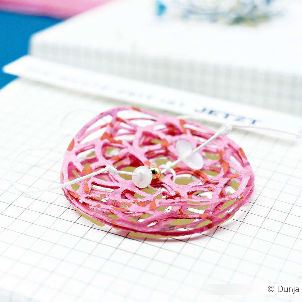 Ein rosafarbener, kreisförmiger, verschlungener Alexandra Renke-Papierausschnitt mit Perlen ruht auf einer weißen Gitterfläche, mit Designpapieren und einem Lineal im Hintergrund.