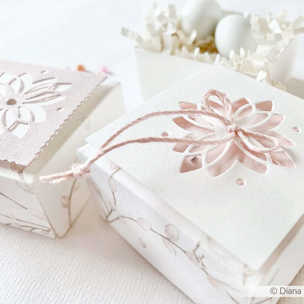 Weiße dekorative Geschenkschachteln mit Blumenausschnitten, die mit der Motivstanze Ornament von Alexandra Renke und Bindfaden gefertigt wurden, stehen neben einer Schachtel, die mit geschreddertem Papier und weißen Eiern gefüllt ist.