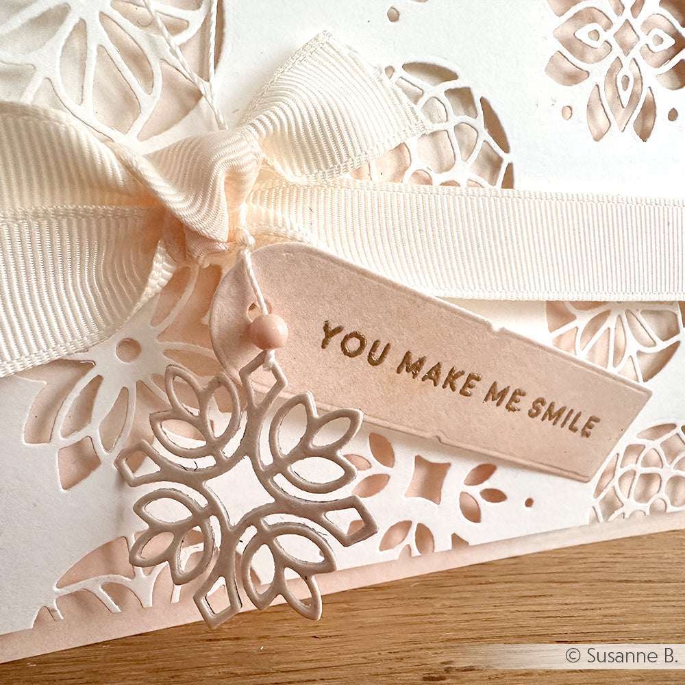 Nahaufnahme einer cremefarbenen Karte mit einer Schleife, Blumenausschnitten, die mit Alexandra Renke Dies für Papierbasteln hergestellt wurden, und einem Anhänger mit der Aufschrift "You make me smile".