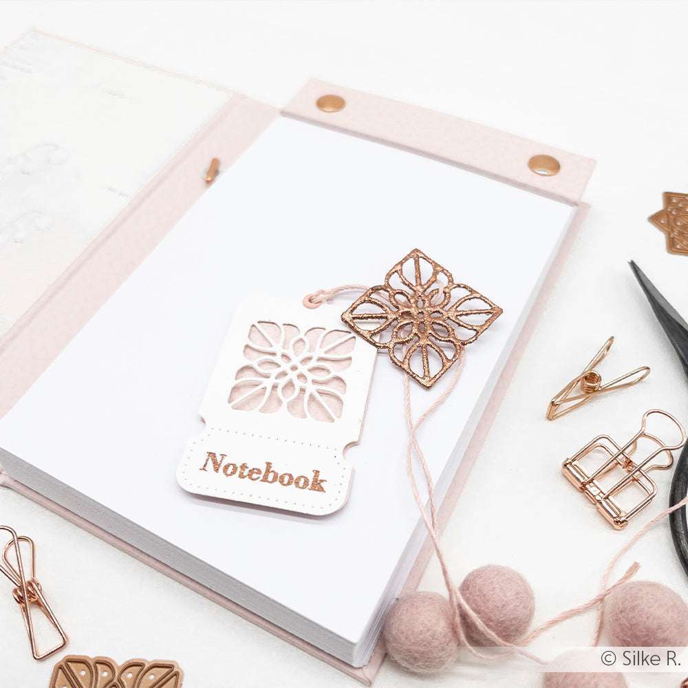 Aufgeschlagenes rosafarbenes Notizbuch von Alexandra Renke mit einem dekorativen Anhänger, goldenen Klammern, Scheren, bunten Designpapieren und Filzkugeln auf einer weißen Oberfläche.