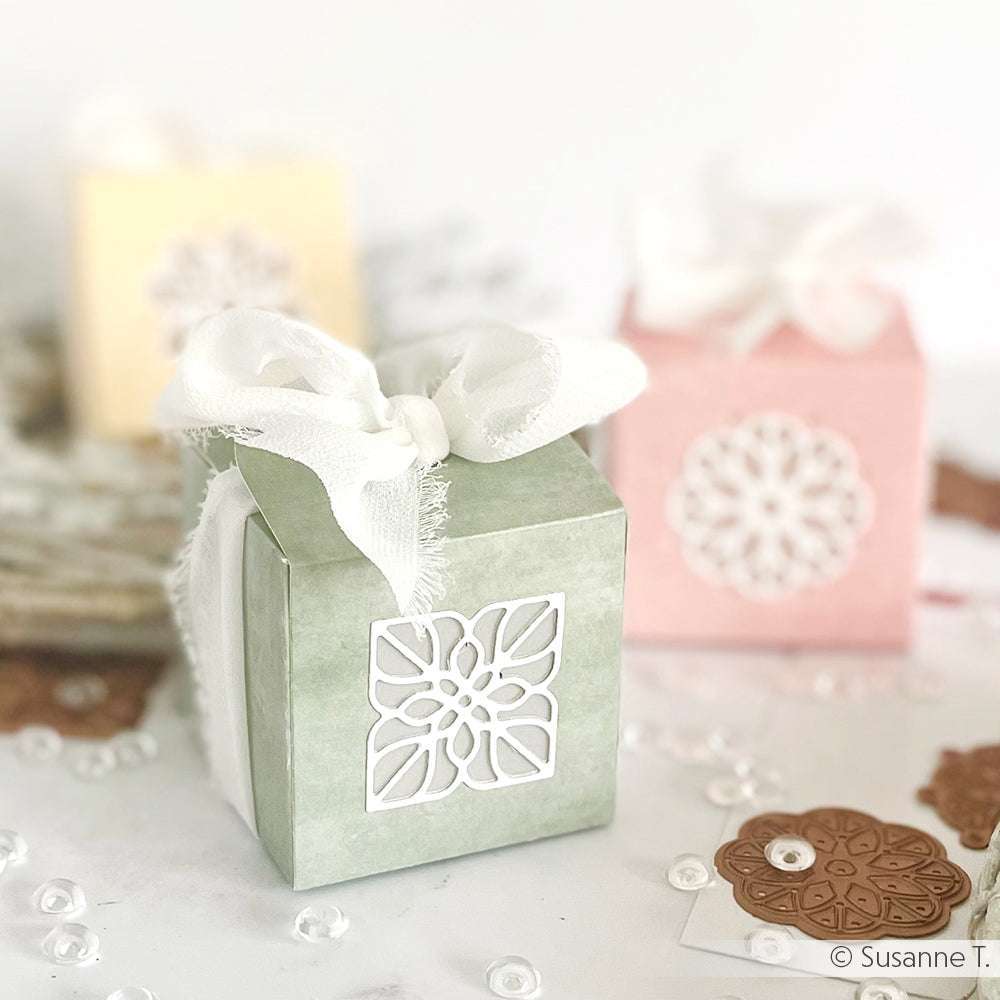 Kleine pastellfarbene Geschenkschachteln mit weißen Bändern und floralen Ausschnitten, die mit den Ornamentstempeln von Alexandra Renke hergestellt wurden, sind von Knöpfen und Pailletten umgeben.