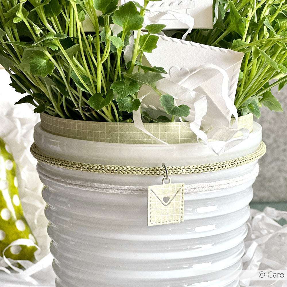 Weißer Keramiktopf mit grünen Blattpflanzen, verziert mit Bändern, einem kleinen quadratischen Anhänger und Akzenten, hergestellt mit Alexandra Renke Designpapieren 200g und Motivstanze Briefe von Herzen aus der Kollektion Stanzsysteme.