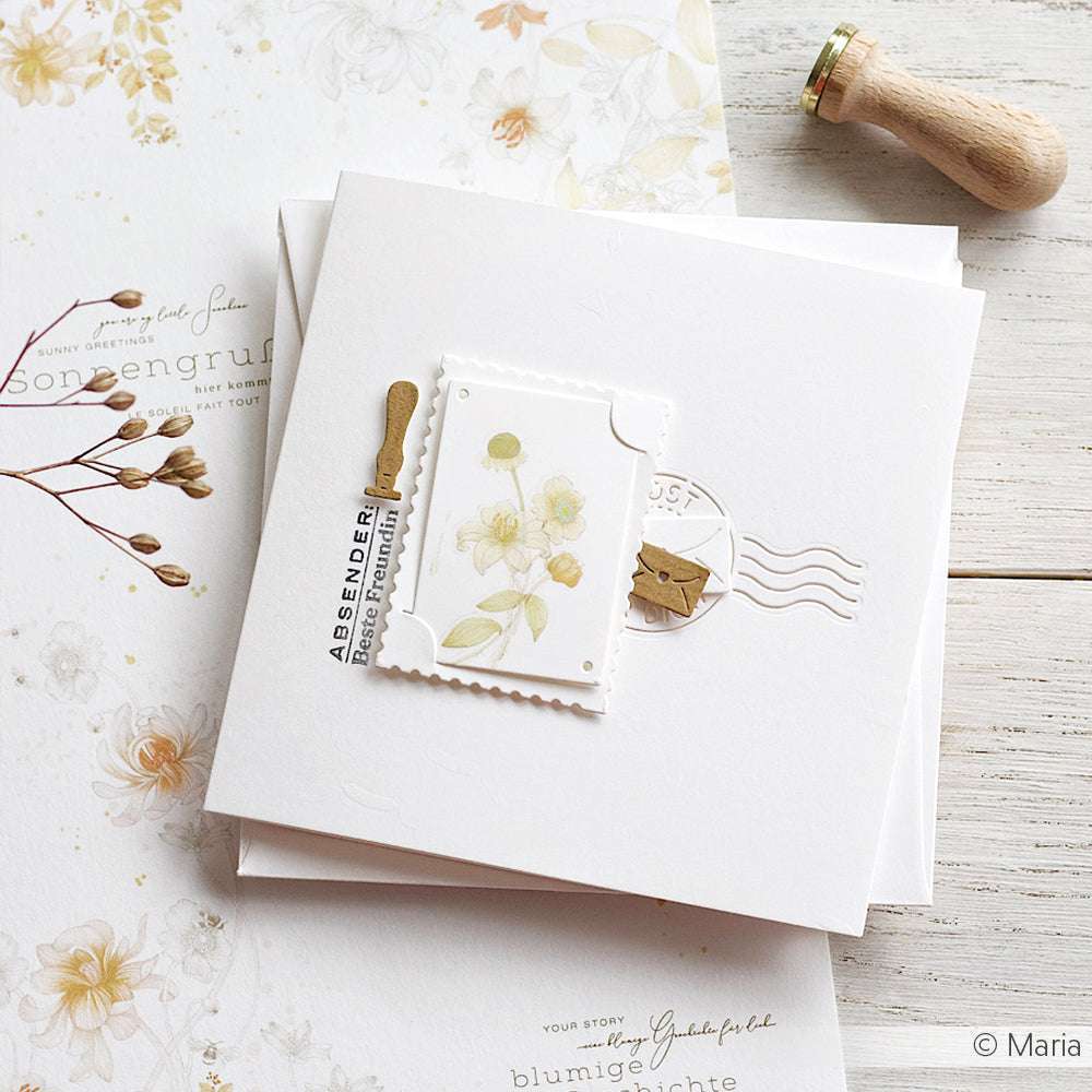 Handgefertigte florale Grußkarte von Alexandra Renke, hergestellt mit Designpapier und einer Steck-Briefmarken-Motivstanze, mit einem Briefmarkenmotiv auf einem weißen Umschlag auf einer Holzoberfläche.