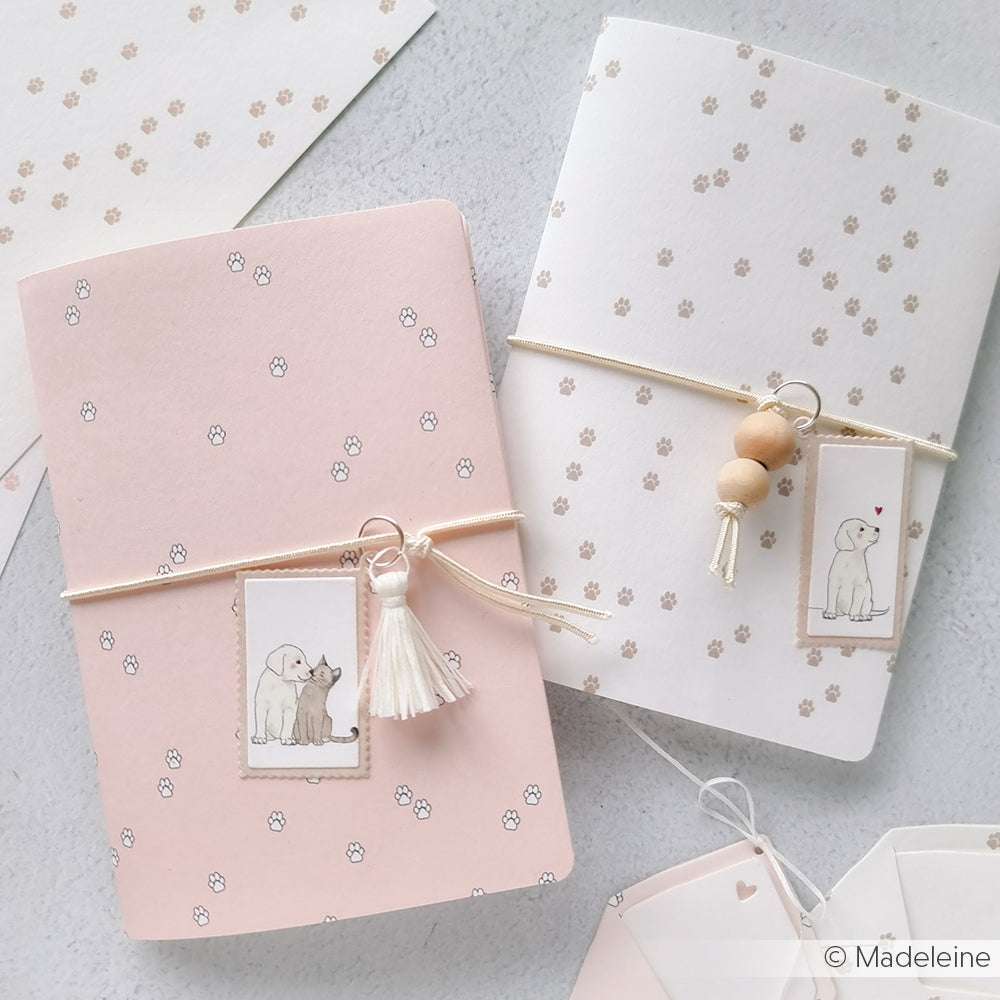 Zwei pastellfarbene Notizbücher von Alexandra Renke mit Tierillustrationen - eines rosa mit einer Katze und einem Hund, das andere weiß mit einem Hund und einer Quaste. Skurrile Designs, perfekt, um sie zusammen mit einer Motivstanze oder einem Briefmarken-Etiketten-Set zu personalisieren.