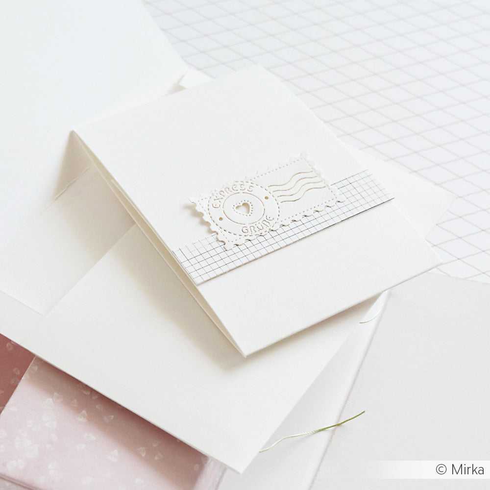 Eine weiße, handgefertigte Karte von Alexandra Renke zeigt ein dekoratives Briefmarkenmotiv, das mit DieCut Systemen auf einem gerasterten Papierstreifen hergestellt wurde und auf weißen Umschlägen liegt.