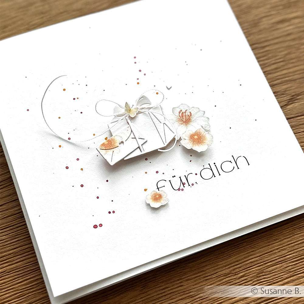 Eine weiße Karte von Alexandra Renke mit 3D-Blumen, einem kleinen Geschenk und dem Schriftzug "für dich" aus Motivstanzen liegt auf einer Holzfläche.
