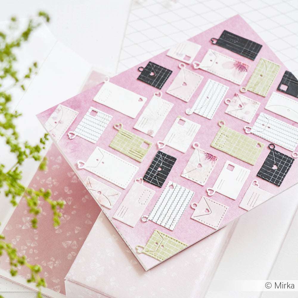 Rosafarbene Karte mit kleinen Papierumschlägen und Notizen, die in der Technik von Alexandra Renke hergestellt wurden, auf einer weißen Gitterfläche neben einer Pflanze platziert.