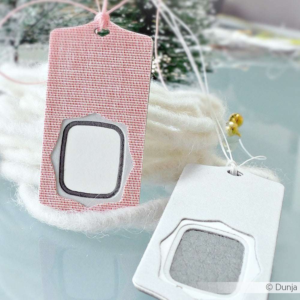 Zwei handgefertigte Alexandra Renke Schichtetikett mit Rahmen Geschenkanhänger, hergestellt mit einer Motivstanze, haben ausgeschnittene Rahmen und Schnur. Sie sind auf einer hellen Fläche mit sanft verschwommener weißer Wolle im Hintergrund ausgestellt.