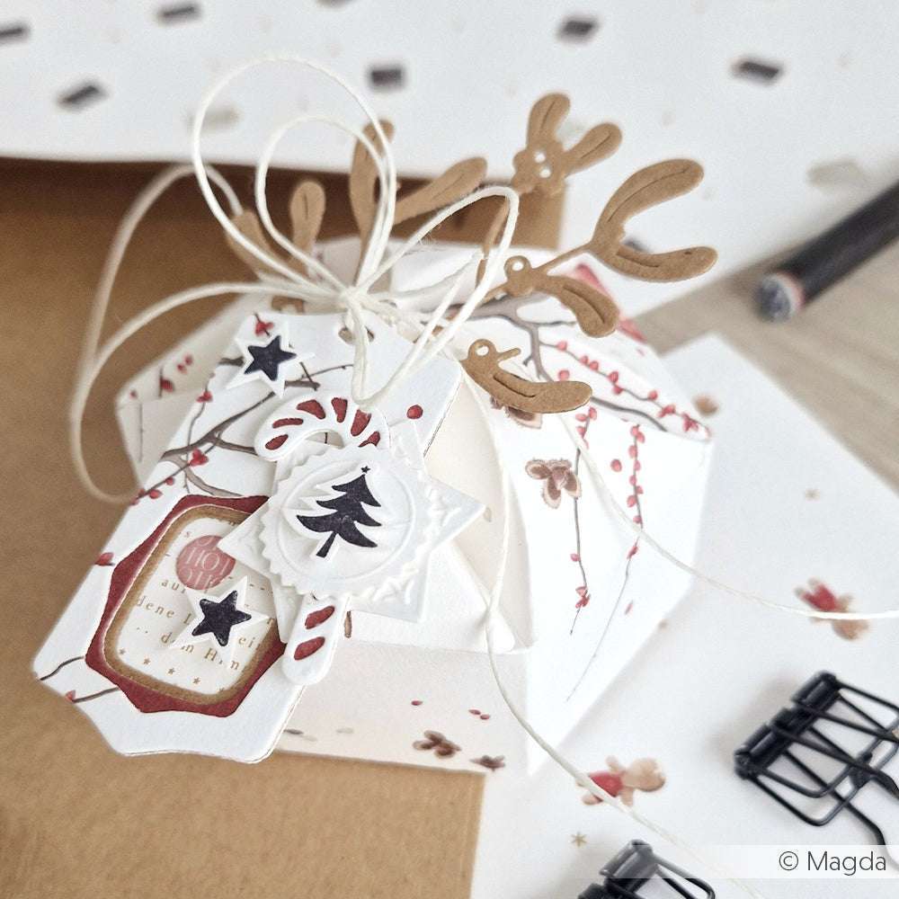 Eine festliche Geschenkschachtel von Alexandra Renke, verziert mit Rentiergeweih, Stern und Baumanhängern, die mit einer Motivstanze ausgeschnitten wurden, sowie mit Schnurakzenten auf einer weißen Oberfläche.