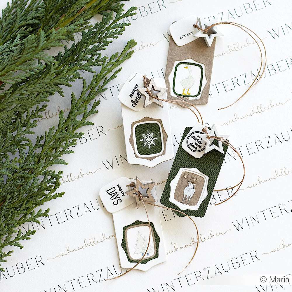 Vier handgefertigte winterliche Geschenkanhänger von Alexandra Renke mit Naturmotiven, hergestellt mit DieCut Systemen und Schichtetikett mit Rahmen, arrangiert auf einer Fläche mit dem Text "WINTERZAUBER".