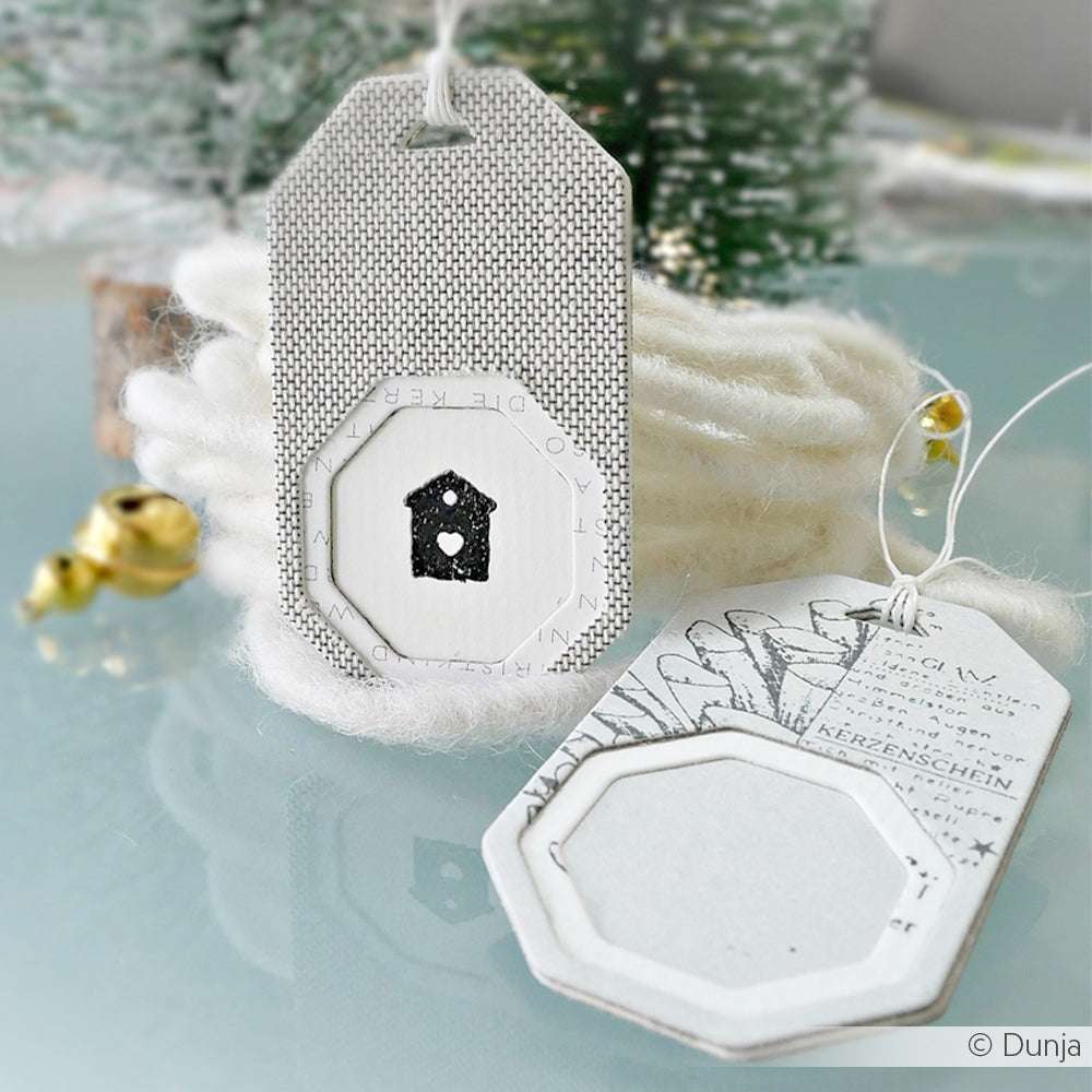 Zwei dekorative weiße Geschenkanhänger von Alexandra Renke, hergestellt mit DieCut Systemen und winterlichen Designpapieren, werden auf einem Tisch mit festlicher Dekoration präsentiert.