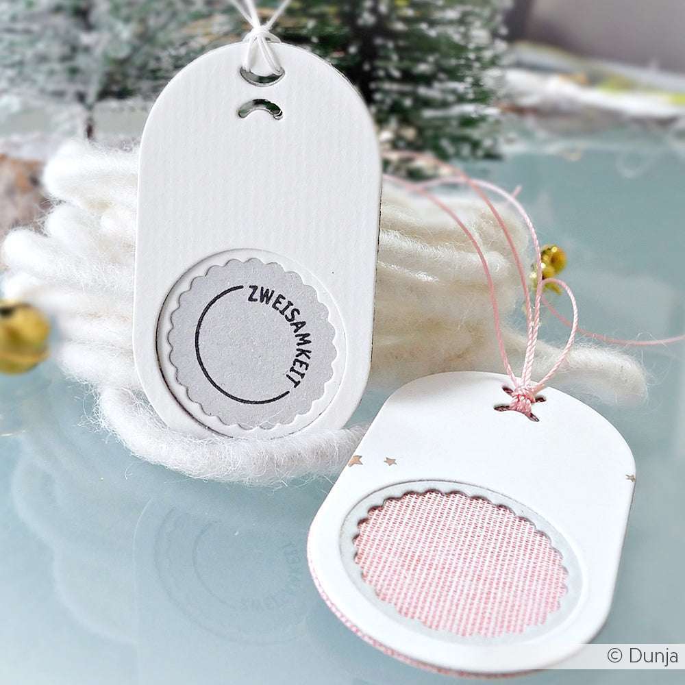 Zwei handgefertigte weiße Geschenkanhänger von Alexandra Renke, die mit DieCut Systemen und Motivstanze Schichtetikett hergestellt wurden, zeigen runde Ausschnitte und den Text "ZWEISAMKEIT" auf einem weichen, winterlichen Hintergrund.