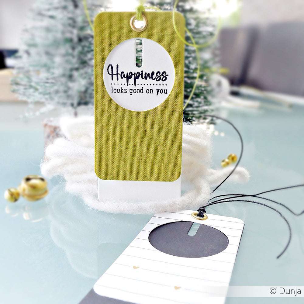 Zwei dekorative Geschenkanhänger von Alexandra Renke, ein grüner mit der Aufschrift "Glück steht dir gut", hergestellt mit den Motivstanze Kombinations-Etikett 2 Stanzformen für Designpapier, ausgestellt auf Weiß in der Nähe eines Baumes.