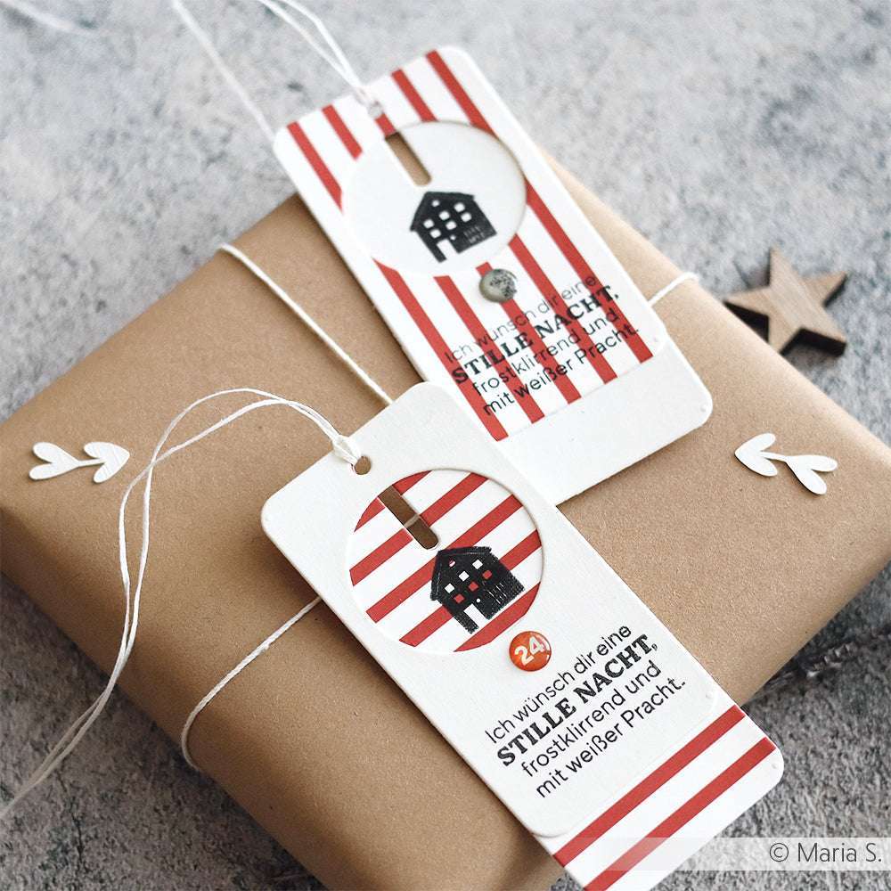 Geschenkverpackung aus braunem Papier mit zwei gestreiften Anhängern, die jeweils ein kleines Haus und einen deutschen Feiertagstext zeigen - gestaltet mit Alexandra Renkes Motivstanze Kombinations-Etikett für einzigartige Formen und veredelt mit festlichen Designpapieren.